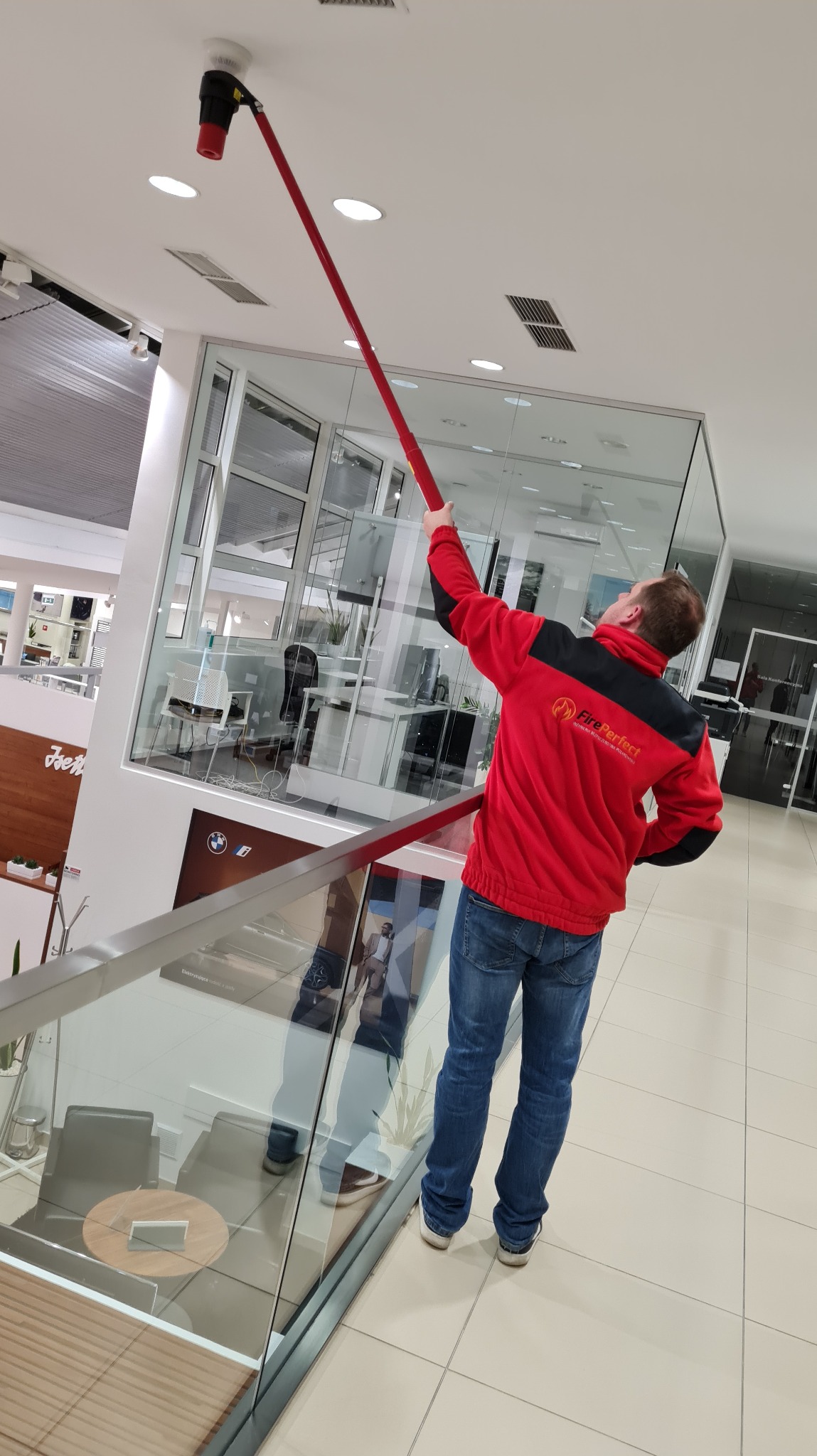 Mężczyzna w czerwonej bluzie z logo Fire Perfect czyści czujnik dymu na suficie w nowoczesnym biurze za pomocą specjalistycznego narzędzia z długim uchwytem.