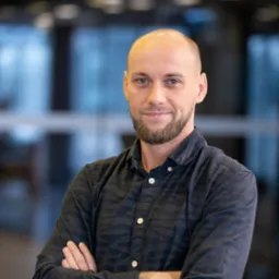 Portret uśmiechniętego mężczyzny z brodą, w ciemnej koszuli w prążki, z założonymi rękami na tle rozmytego biura.