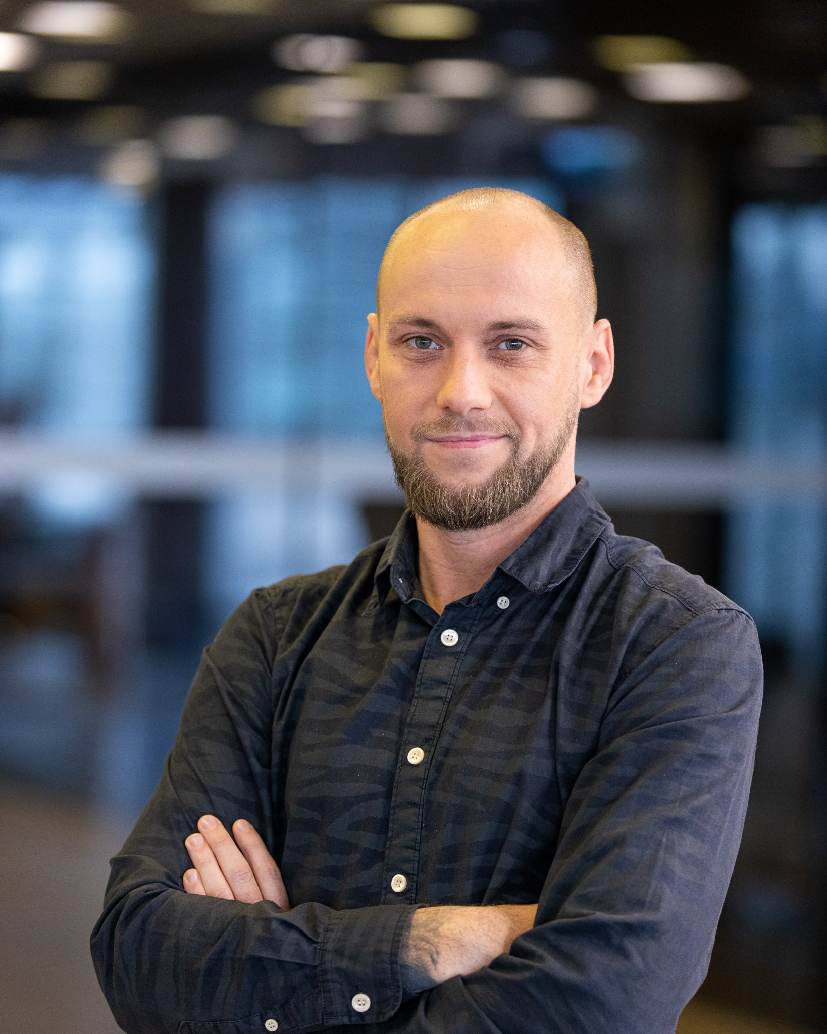 Portret uśmiechniętego mężczyzny z brodą, w ciemnej koszuli w prążki, z założonymi rękami na tle rozmytego biura.