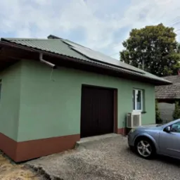 Świeżo pomalowany budynek gospodarczy z brązowym pasem u dołu, zieloną elewacją, brązową bramą garażową i panelami słonecznymi na dachu, obok zaparkowany srebrny samoch&oacute;d.