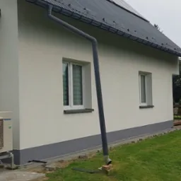 Ściana budynku z ociepleniem, nową elewacją w kolorze kremowym, szarą podmurówką i orynnowaniem; na dachu panele słoneczne. Teren wokół budynku z fragmentami kostki brukowej i trawnikiem.