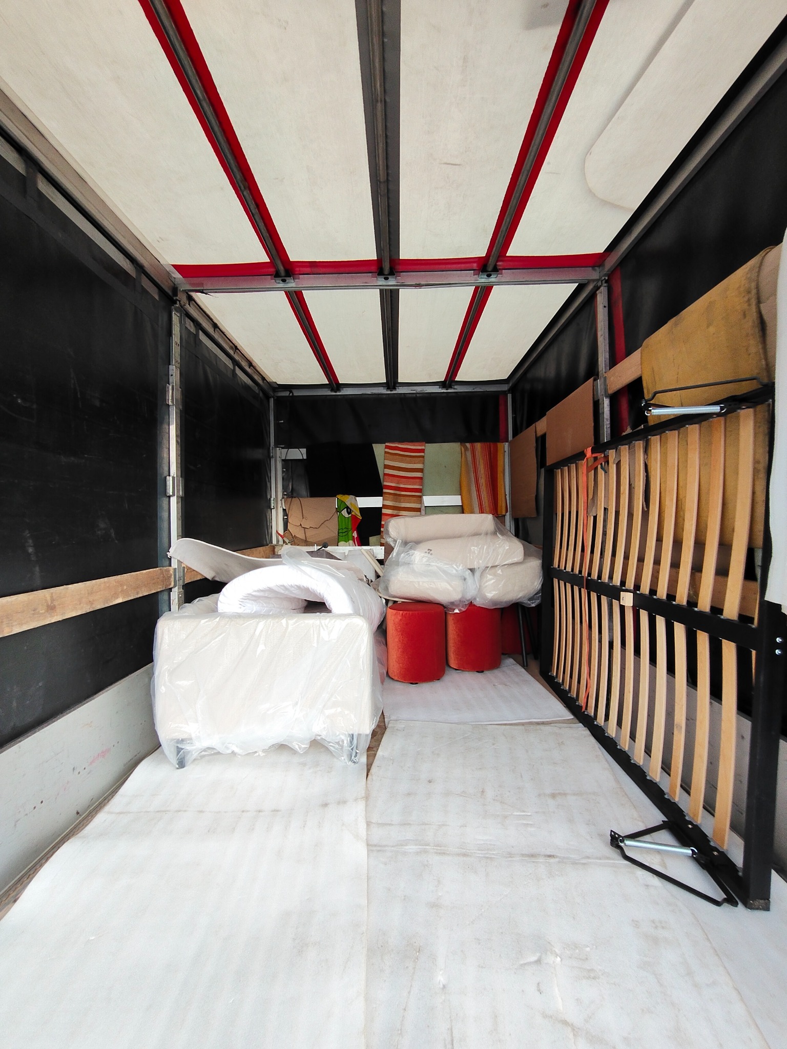 Wnętrze busa transportowego z meblami: łóżko ze stelażem, pufy, materace i inne przedmioty zabezpieczone folią, gotowe do przewozu. Widok z tyłu pojazdu.