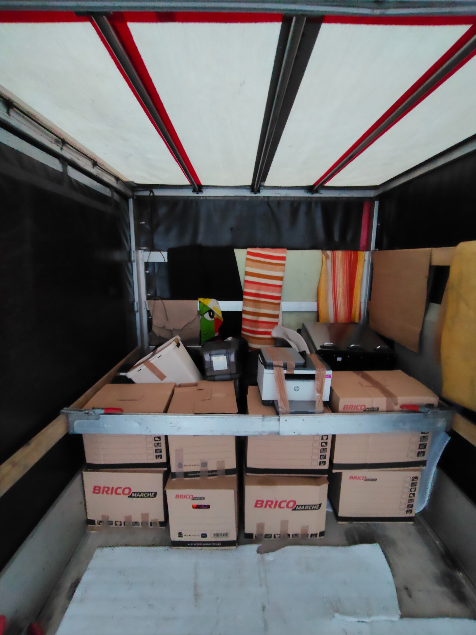 Wnętrze busa wypełnione kartonami Brico Marche i różnymi przedmiotami, gotowe do transportu. Widoczne zabezpieczenia ładunku i oświetlenie wewnętrzne.