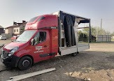 Czerwony bus z otwartą platformą załadunkową, na której zabezpieczone są czarne, cylindryczne obiekty, prawdopodobnie butle lub zbiorniki, widoczne na tle szarego nieba i zabudowań.