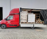 Czerwony bus dostawczy z otwartą przestrzenią ładunkową, wypełnioną paletami z zapakowanymi kartonami i innymi paczkami, widok z boku na tle szarego budynku.