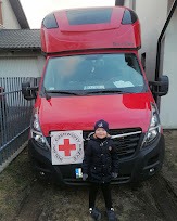 Czerwony bus z wysoką kabiną, z przodu widoczna dziewczynka trzymająca kartkę z czerwonym krzyżem i napisem 'CHARYTATYWNY BUS'.