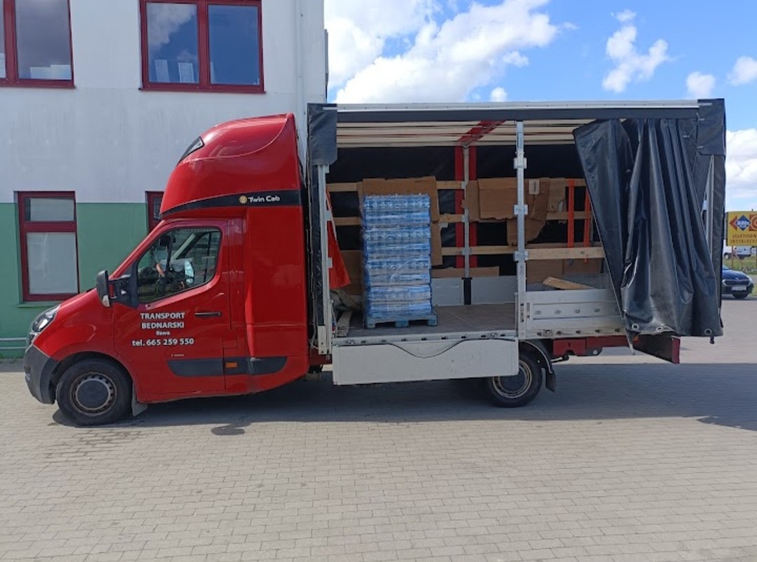 Czerwony bus transportowy z otwartą przestrzenią ładunkową, w której widoczne są palety z wodą butelkowaną i kartony, zaparkowany na brukowanym placu.