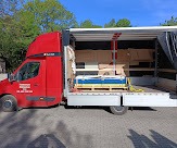 Czerwony samochód ciężarowy z otwartą przestrzenią ładunkową, wypełnioną paletami z zapakowanymi kartonami i zabezpieczone pasami transportowymi, widoczne drzewa w tle.