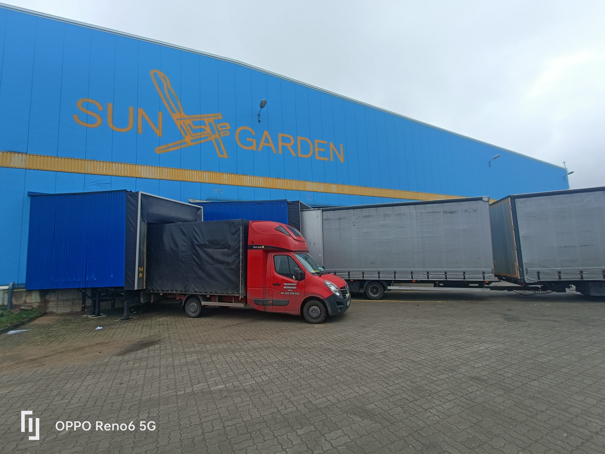 Czerwony bus z czarną plandeką firmy Transport Bednarski zaparkowany przy niebieskim magazynie Sun Garden, obok zaparkowane szare naczepy tirów.