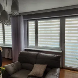 Beżowo-białe, poziome przesłony okienne w nowoczesnym salonie z szarą sofą i wzorzystymi zasłonami po bokach okien.