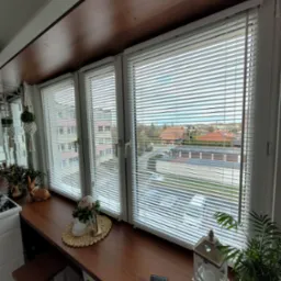 Trzy okna z aluminiowymi żaluzjami poziomymi, widok zza blatu z dekoracjami i roślinami, widoczne budynki za oknem.