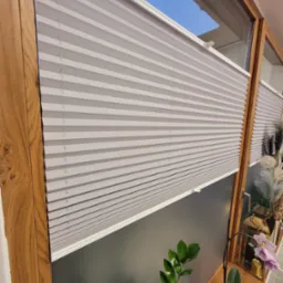 Szare plisy okienne zamontowane w drewnianej ramie okiennej, widok z bliska na strukturę materiału i detale montażu, z roślinami doniczkowymi na parapecie.