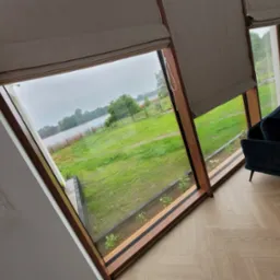 Beżowe rolety rzymskie zamontowane w oknach z drewnianymi ramami, widok na jezioro i trawnik, fragment ciemnoniebieskiej sofy i kosz z wikliny.