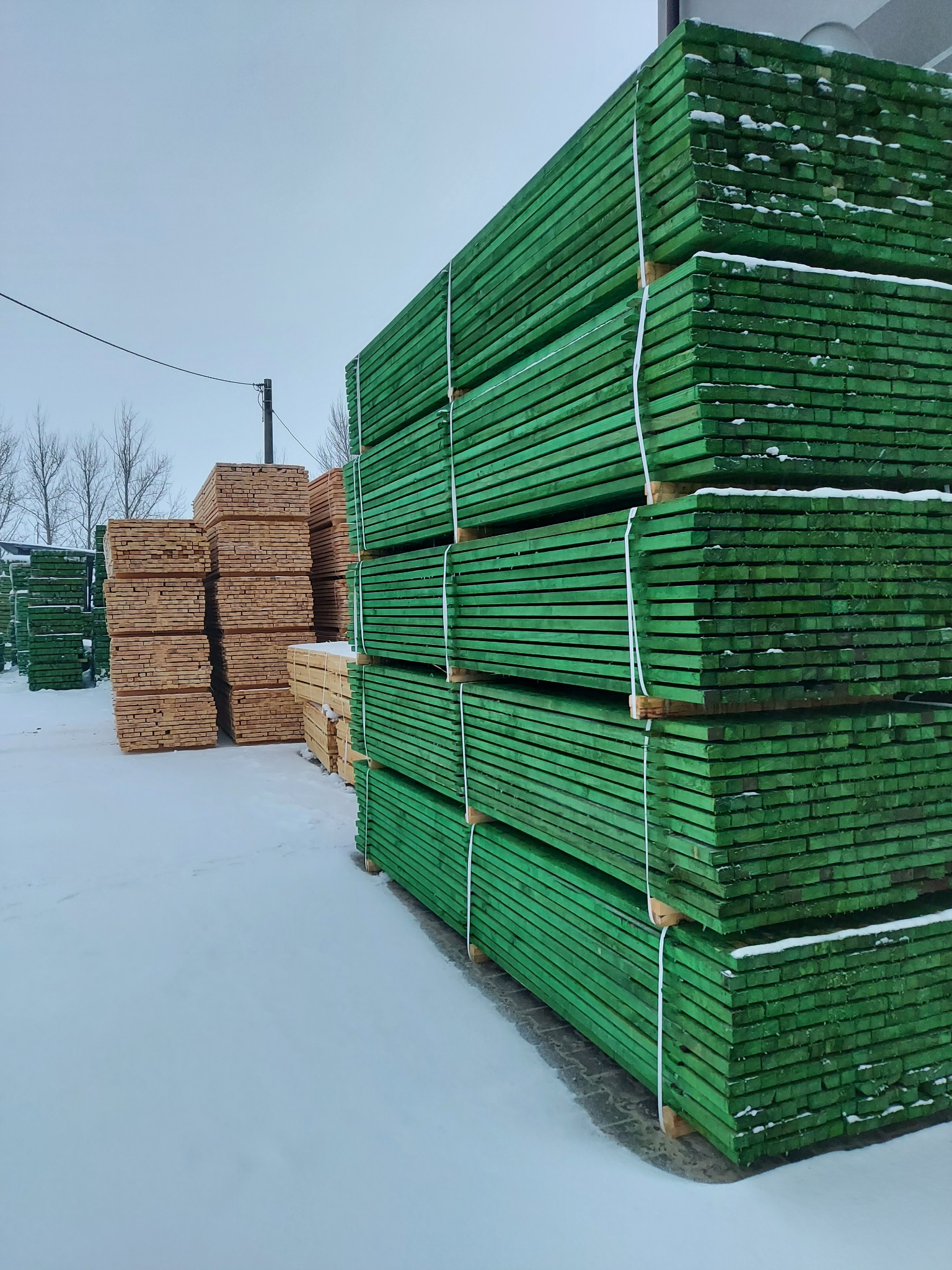 Sztaple impregnowanej na zielono tarcicy budowlanej, zabezpieczone taśmami, ułożone na śniegu; w tle widoczne inne, nieimpregnowane stosy drewna.
