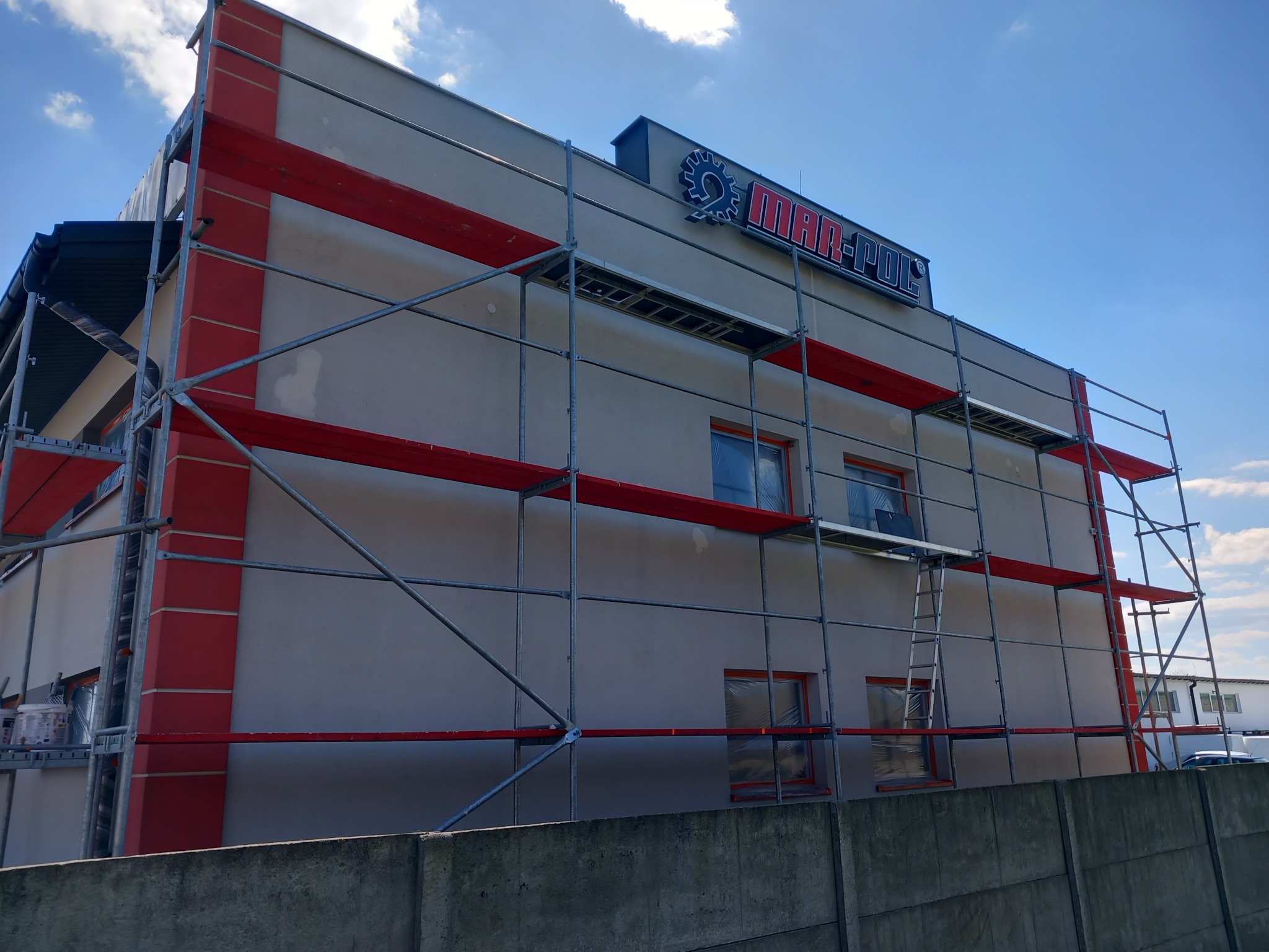 Budynek w trakcie prac elewacyjnych, widoczne rusztowania, zabezpieczone okna folią i logo firmy na dachu.