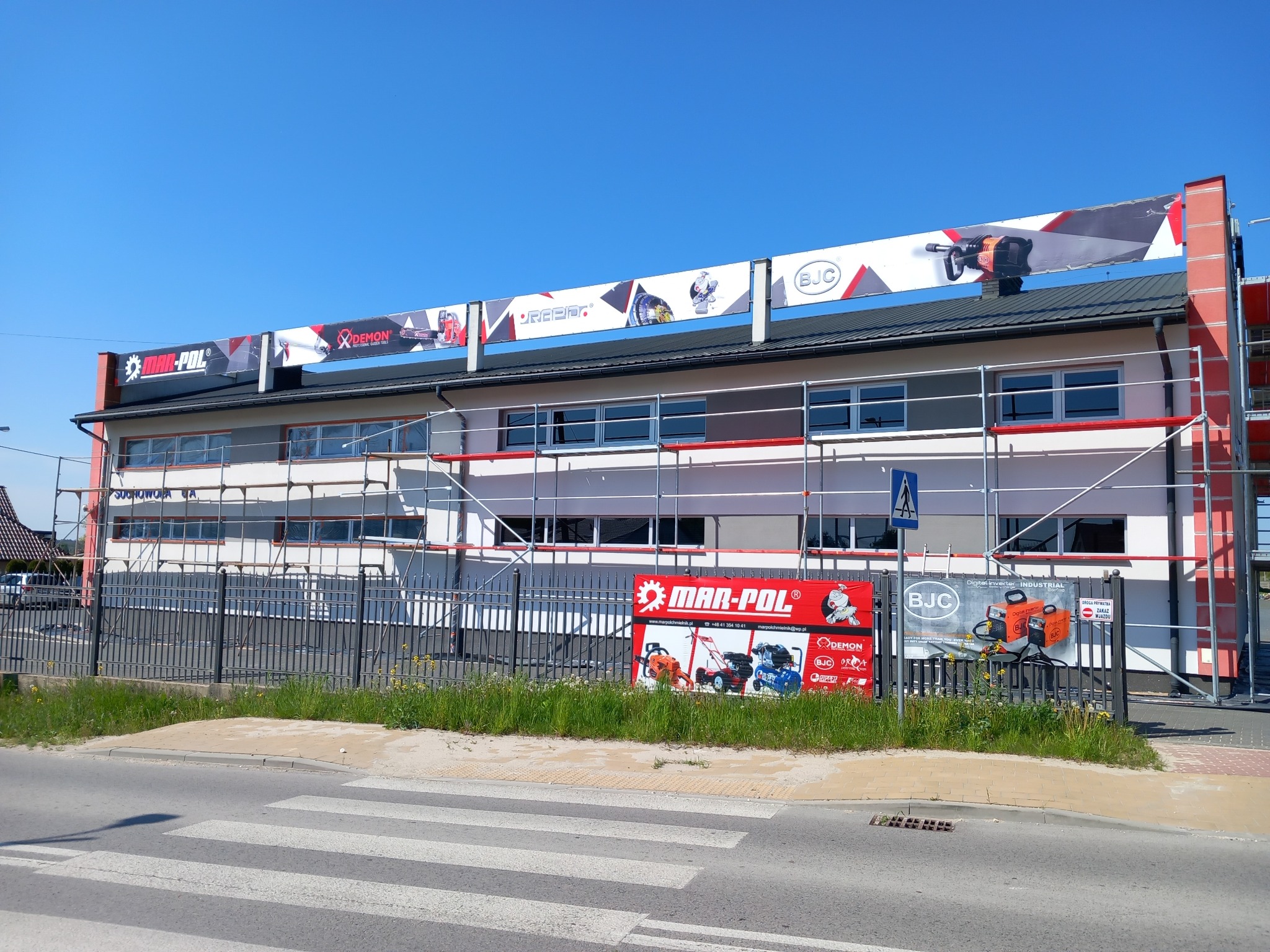 Budynek firmy w Pierzchnicy w trakcie prac elewacyjnych, widoczne rusztowania i banery reklamowe narzędzi. Na ogrodzeniu umieszczone dodatkowe reklamy firmy.