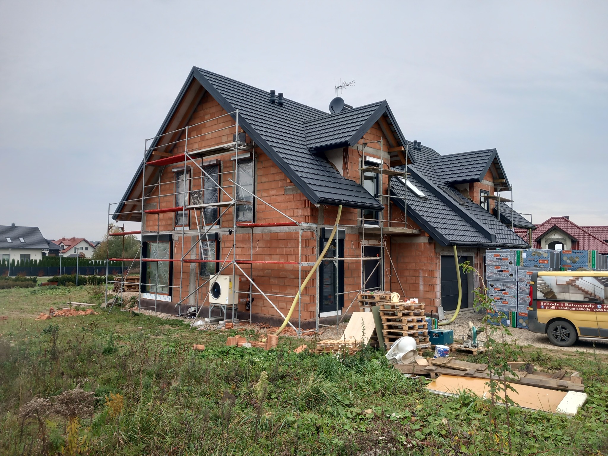Surowy stan domu jednorodzinnego z czerwonej cegły, z czarną dachówką, w trakcie budowy, z widocznym rusztowaniem i materiałami budowlanymi na posesji, na tle pochmurnego nieba.