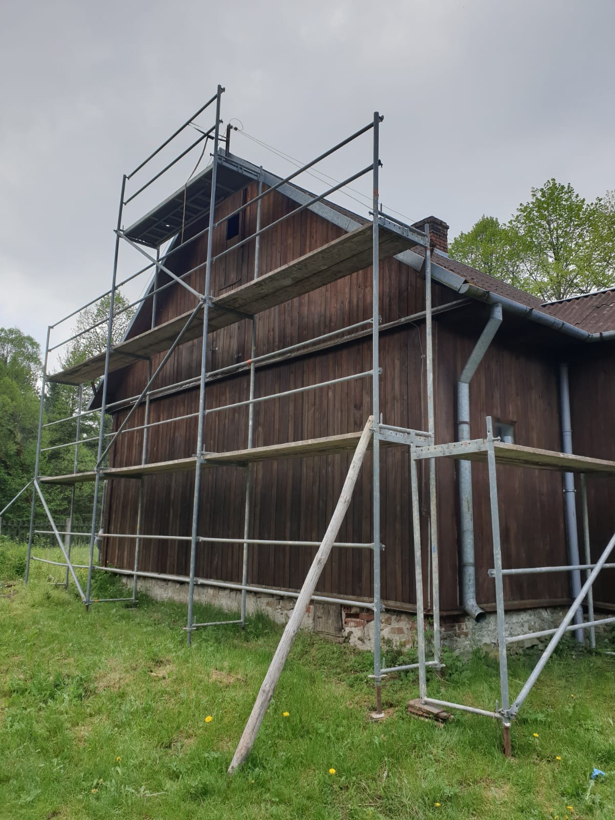 Drewniany dom z ciemnego drewna otoczony rusztowaniem na tle pochmurnego nieba, widoczne rynny i komin