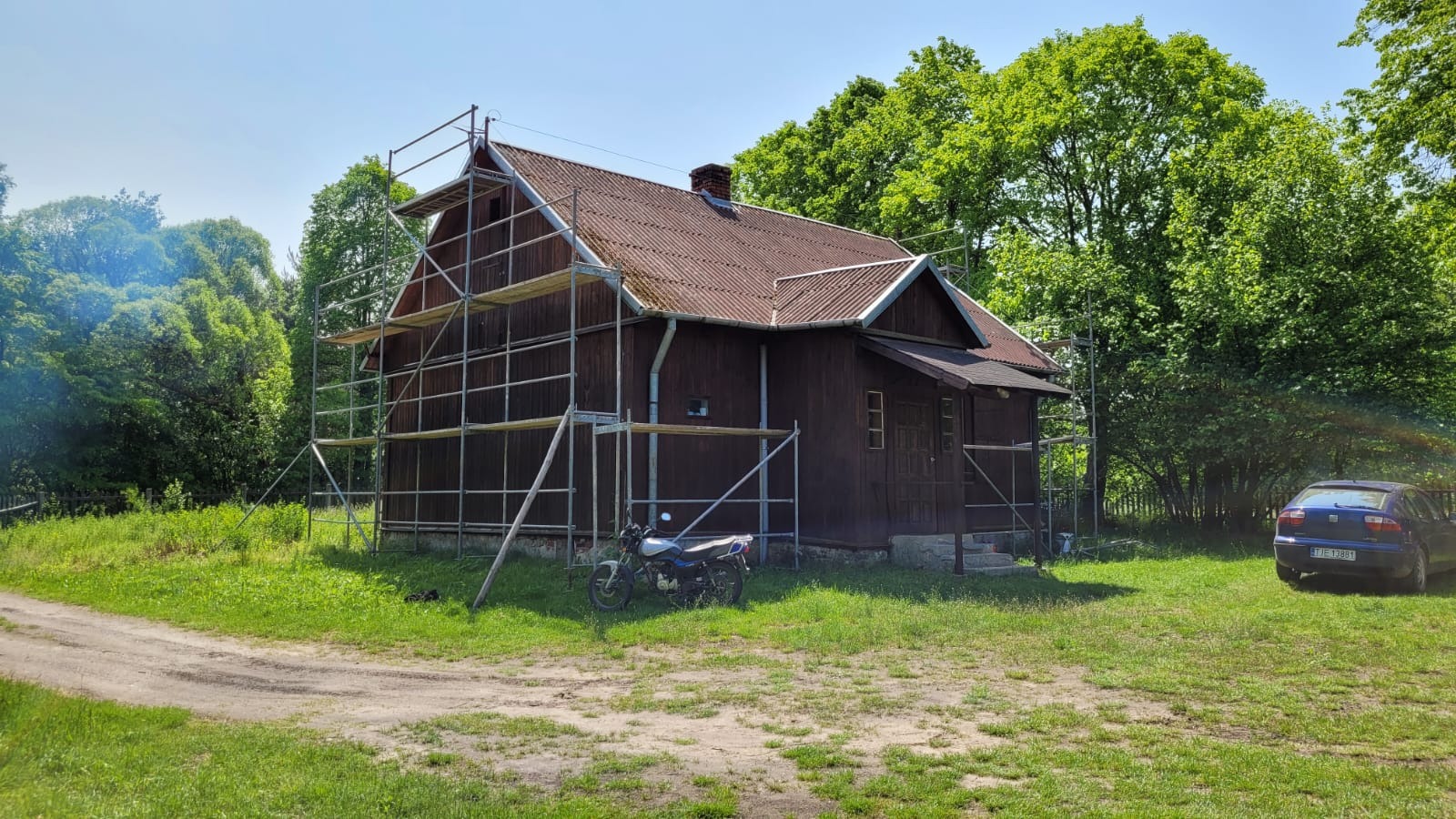 Rustykalny, ciemnobrązowy dom z rusztowaniem na tle zieleni, obok zaparkowany motocykl i samochód.