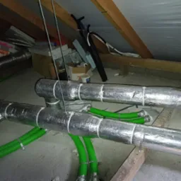 Izolowane, srebrną folią przewody wentylacyjne z zielonymi rurami w przestrzeni poddasza, zamocowane do drewnianej konstrukcji dachu, z widocznymi elementami instalacji i materiałami budowlanymi...