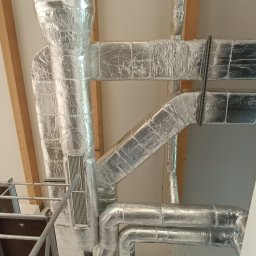 Marcin Oleksyk - System kanałów wentylacyjnych i rur, owiniętych srebrną folią izolacyjną, zainstalowany pod sufitem z widocznymi drewnianymi belkami konstrukcyjnymi, widziany z perspektywy rusztowania.
