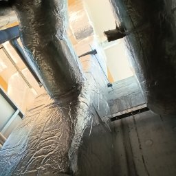 Marcin Oleksyk - Izolowane kanały wentylacyjne, owinięte folią aluminiową, biegną pionowo wzdłuż betonowej ściany, widoczne elementy montażowe i konstrukcja sufitu.