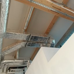 Marcin Oleksyk - Widok z dołu na system kanałów wentylacyjnych owiniętych folią aluminiową, zamontowanych pod sufitem z widocznymi drewnianymi belkami konstrukcyjnymi i metalowymi szynami montażowymi.