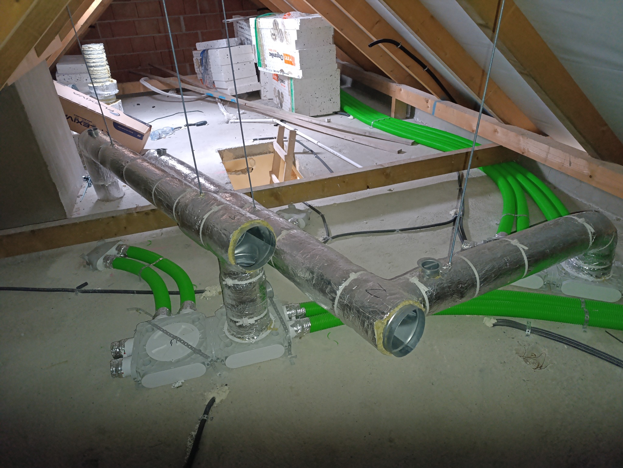 Szare rury wentylacyjne owinięte folią aluminiową, podwieszone na stalowych linkach w surowym wnętrzu poddasza z widoczną drewnianą konstrukcją dachu i zielonymi, elastycznymi przewodami...