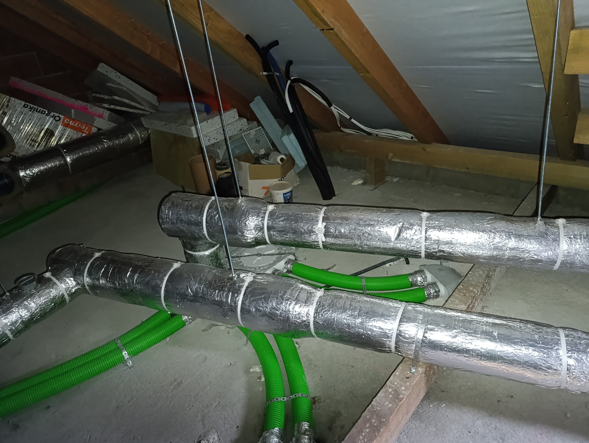 Izolowane, srebrną folią przewody wentylacyjne z zielonymi rurami w przestrzeni poddasza, zamocowane do drewnianej konstrukcji dachu, z widocznymi elementami instalacji i materiałami budowlanymi...