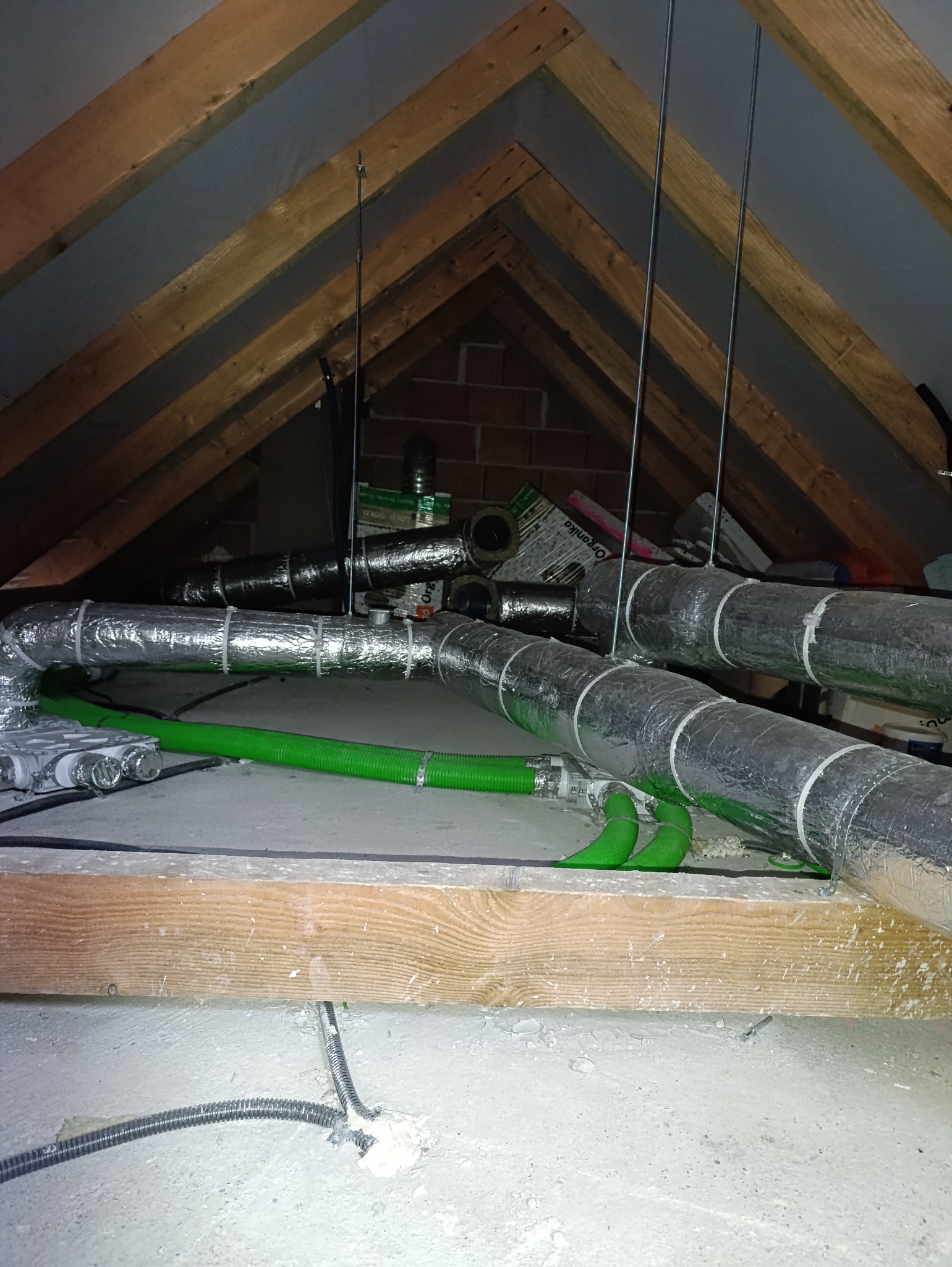Instalacja wentylacyjna z aluminiowymi rurami i zielonymi przewodami elastycznymi w przestrzeni poddasza, widoczne drewniane elementy konstrukcji dachu i ceglana ściana w tle.