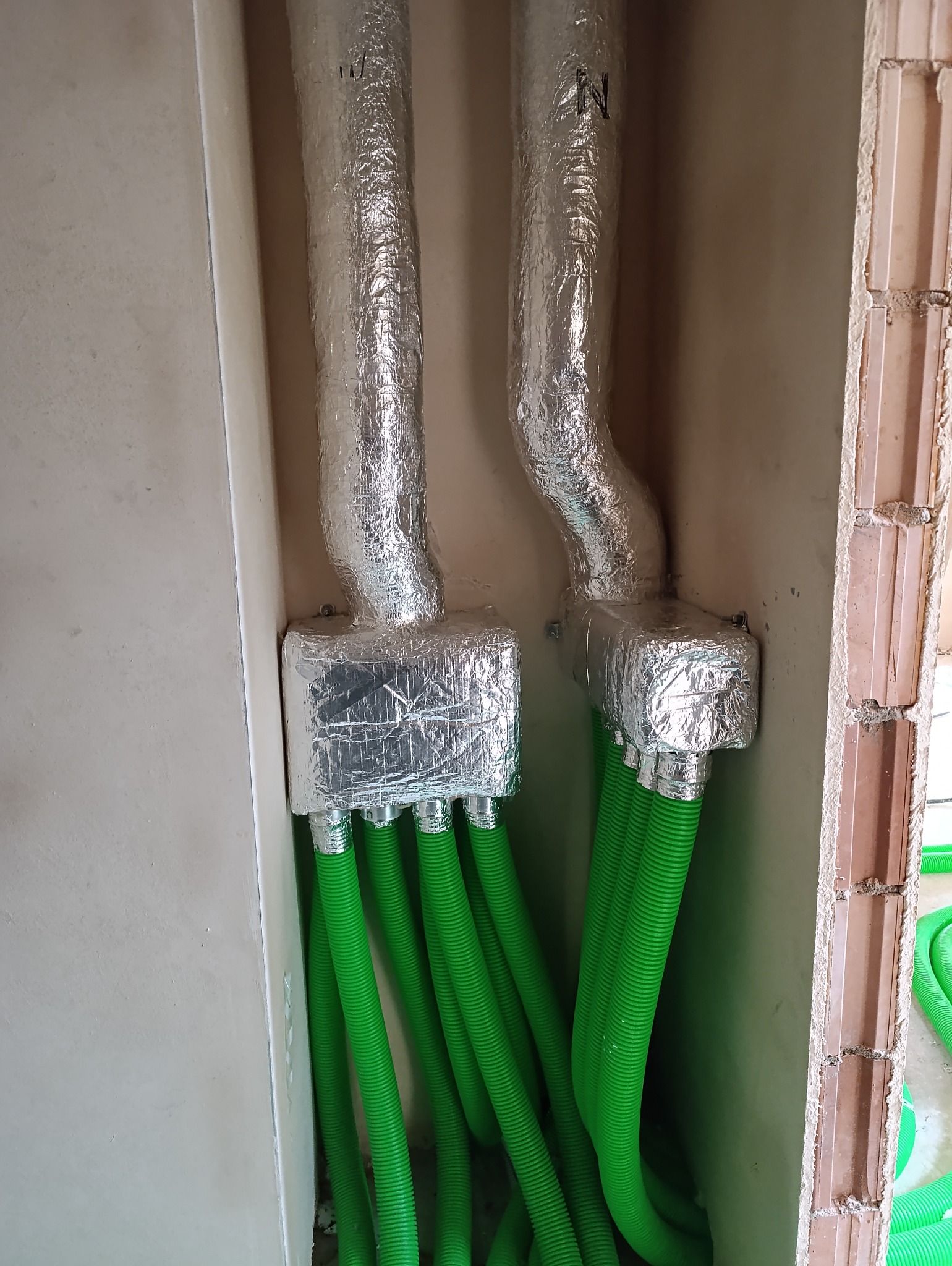 Dwa przewody wentylacyjne owinięte folią aluminiową, z podłączonymi zielonymi rurami karbowanymi, zamocowane w ścianie z cegły.