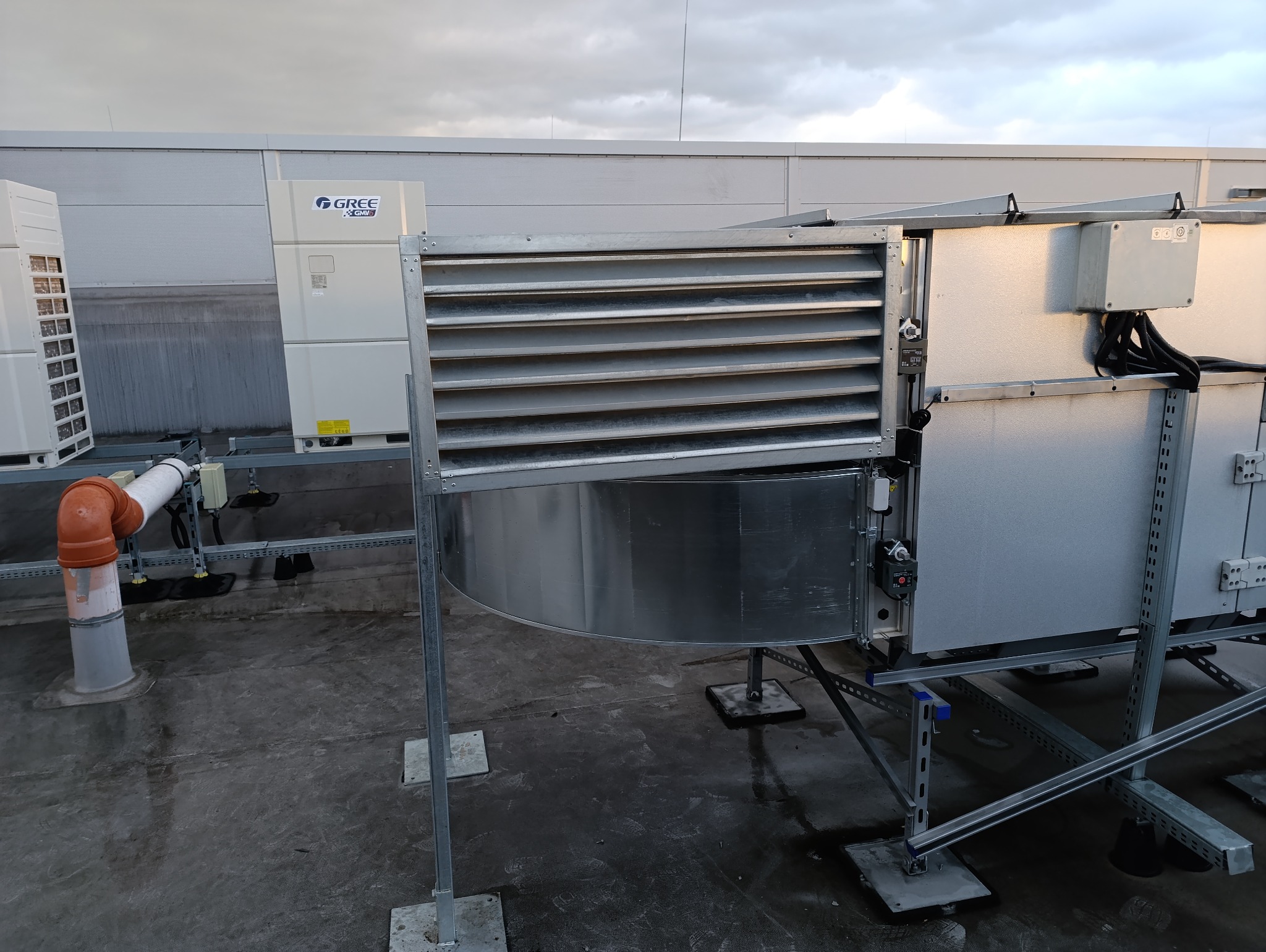 Instalacja wentylacyjna na dachu budynku z widocznymi kanałami wentylacyjnymi, kratkami i rurami odprowadzającymi skropliny, z logo GREE na jednym z urządzeń.