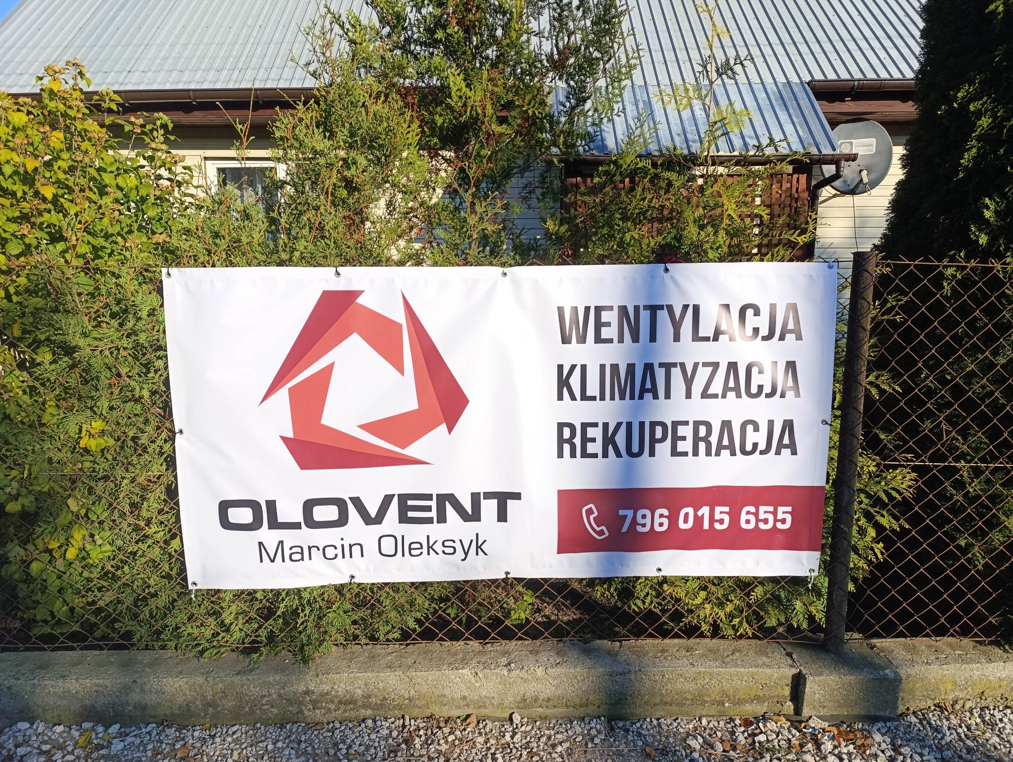 Baner reklamowy firmy OLOVENT z Tarnowa, oferującej wentylację, klimatyzację i rekuperację, zawieszony na ogrodzeniu z siatki, widoczny numer telefonu.