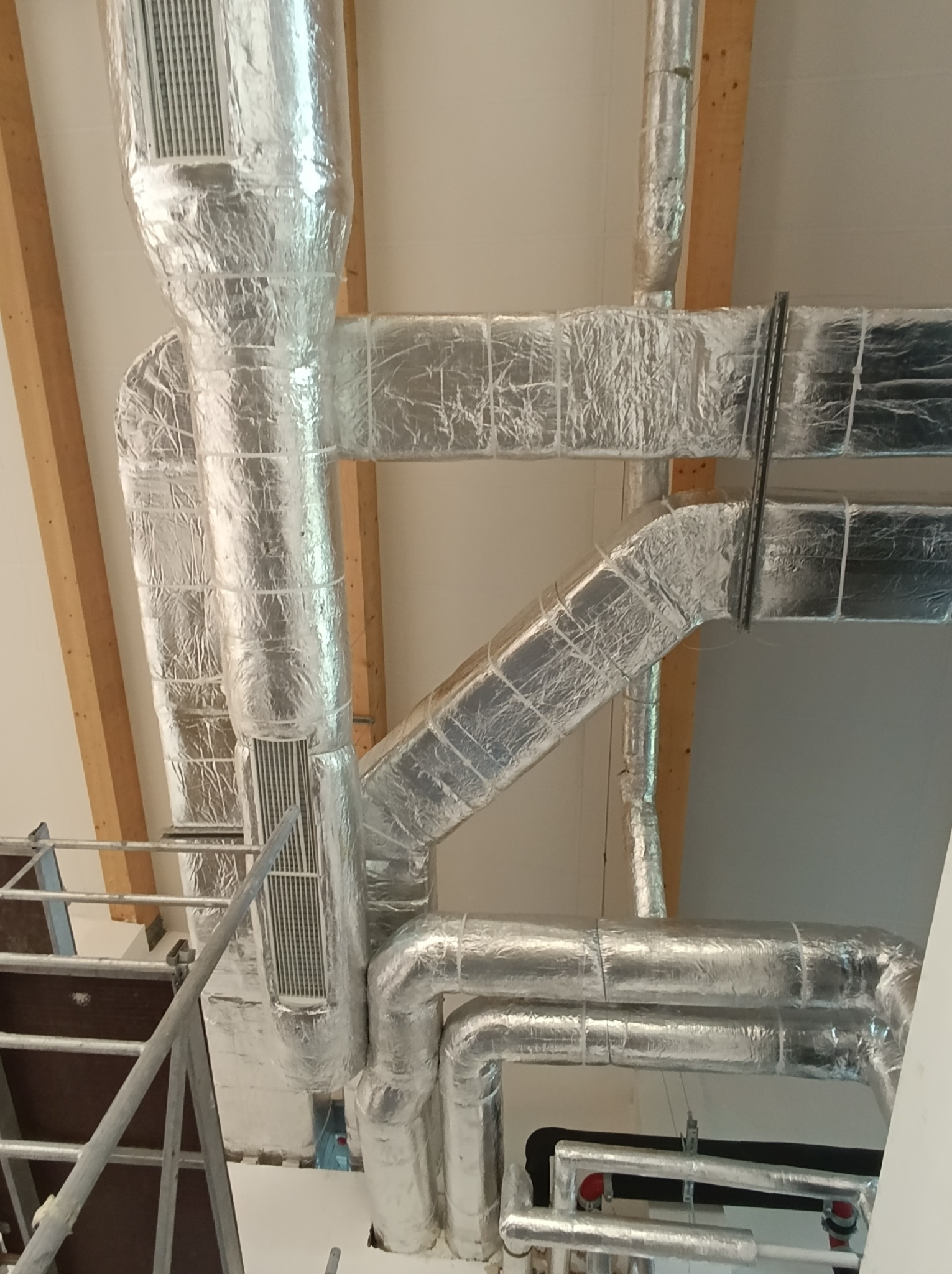 System kanałów wentylacyjnych i rur, owiniętych srebrną folią izolacyjną, zainstalowany pod sufitem z widocznymi drewnianymi belkami konstrukcyjnymi, widziany z perspektywy rusztowania.