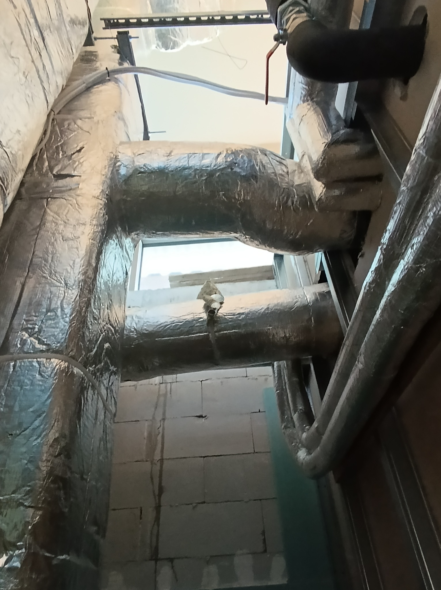 Srebrne, izolowane rury wentylacyjne w surowym wnętrzu z bloków betonowych, widoczne elementy instalacji i zawór z czerwoną rączką.