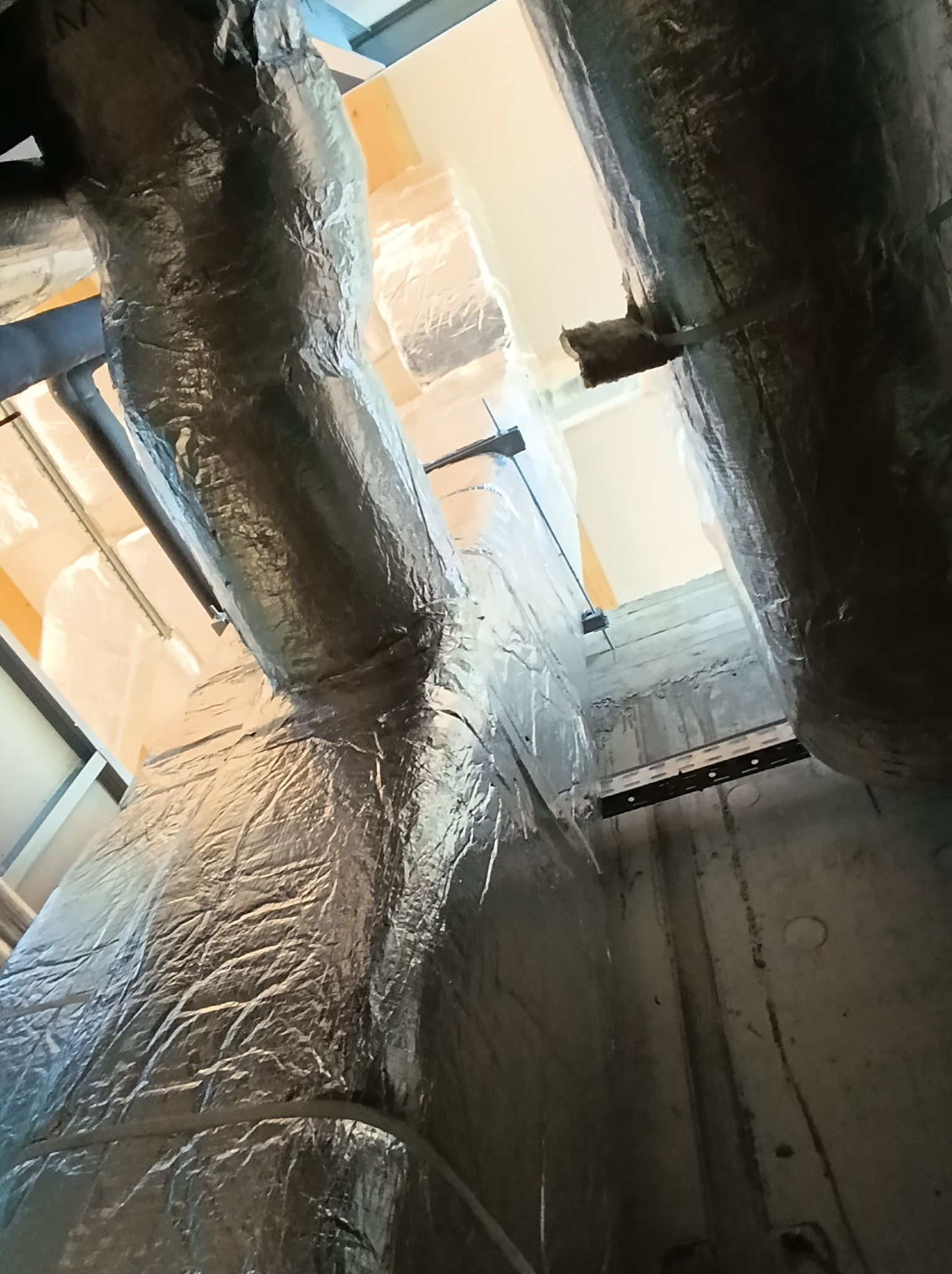 Izolowane kanały wentylacyjne, owinięte folią aluminiową, biegną pionowo wzdłuż betonowej ściany, widoczne elementy montażowe i konstrukcja sufitu.