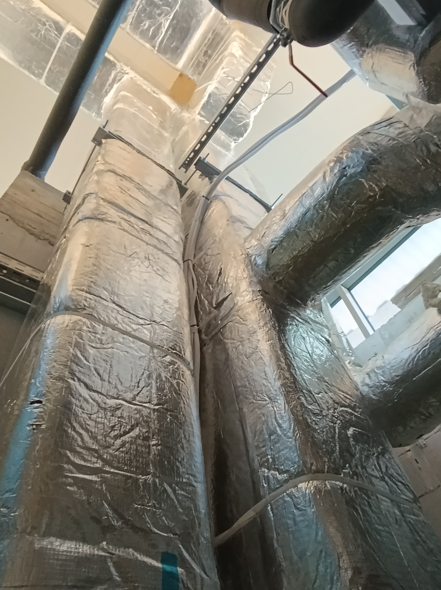 Izolowane kanały wentylacyjne w budynku, widoczne metalowe wsporniki i rury, instalacja w trakcie realizacji.
