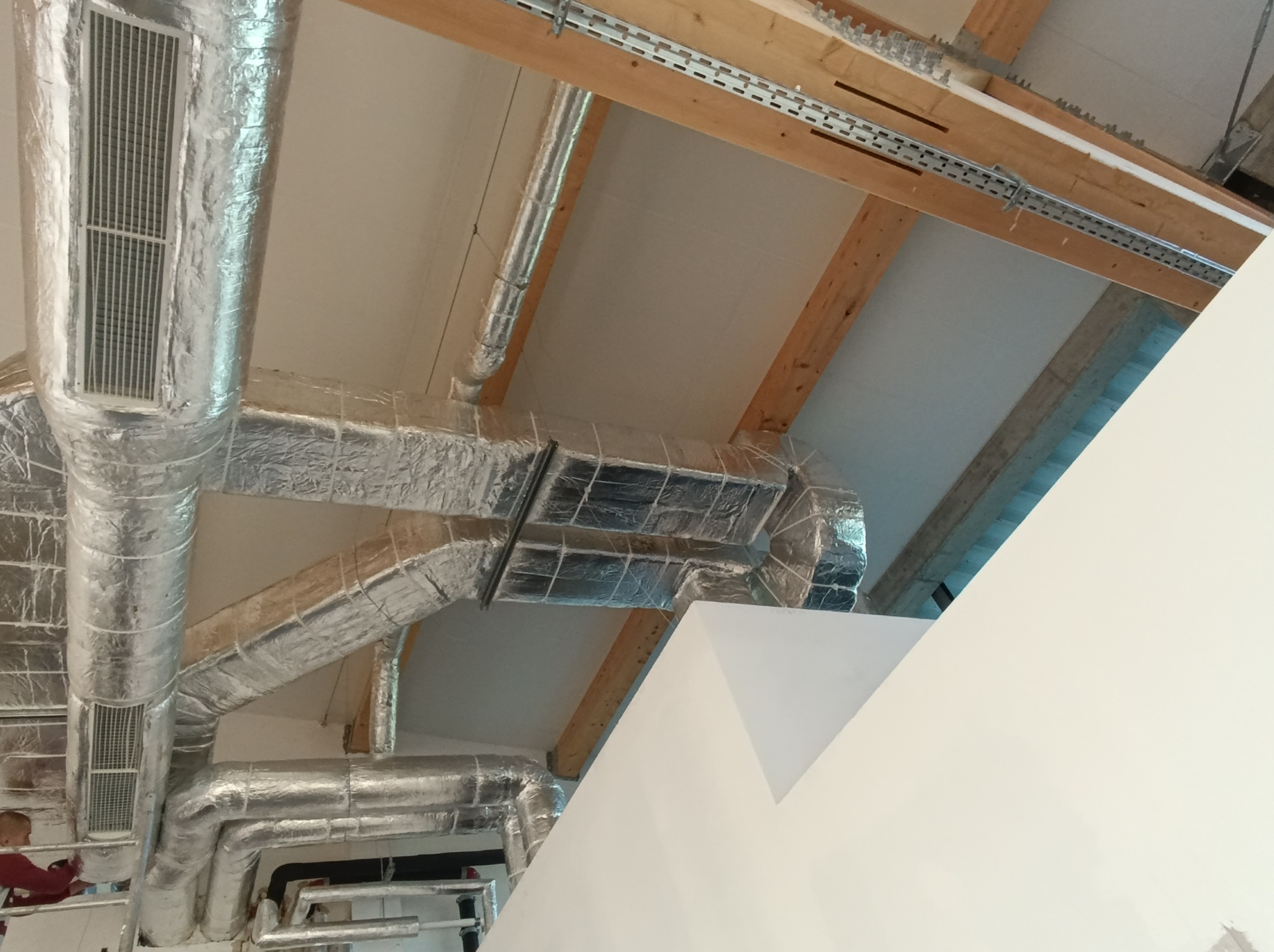 Widok z dołu na system kanałów wentylacyjnych owiniętych folią aluminiową, zamontowanych pod sufitem z widocznymi drewnianymi belkami konstrukcyjnymi i metalowymi szynami montażowymi.