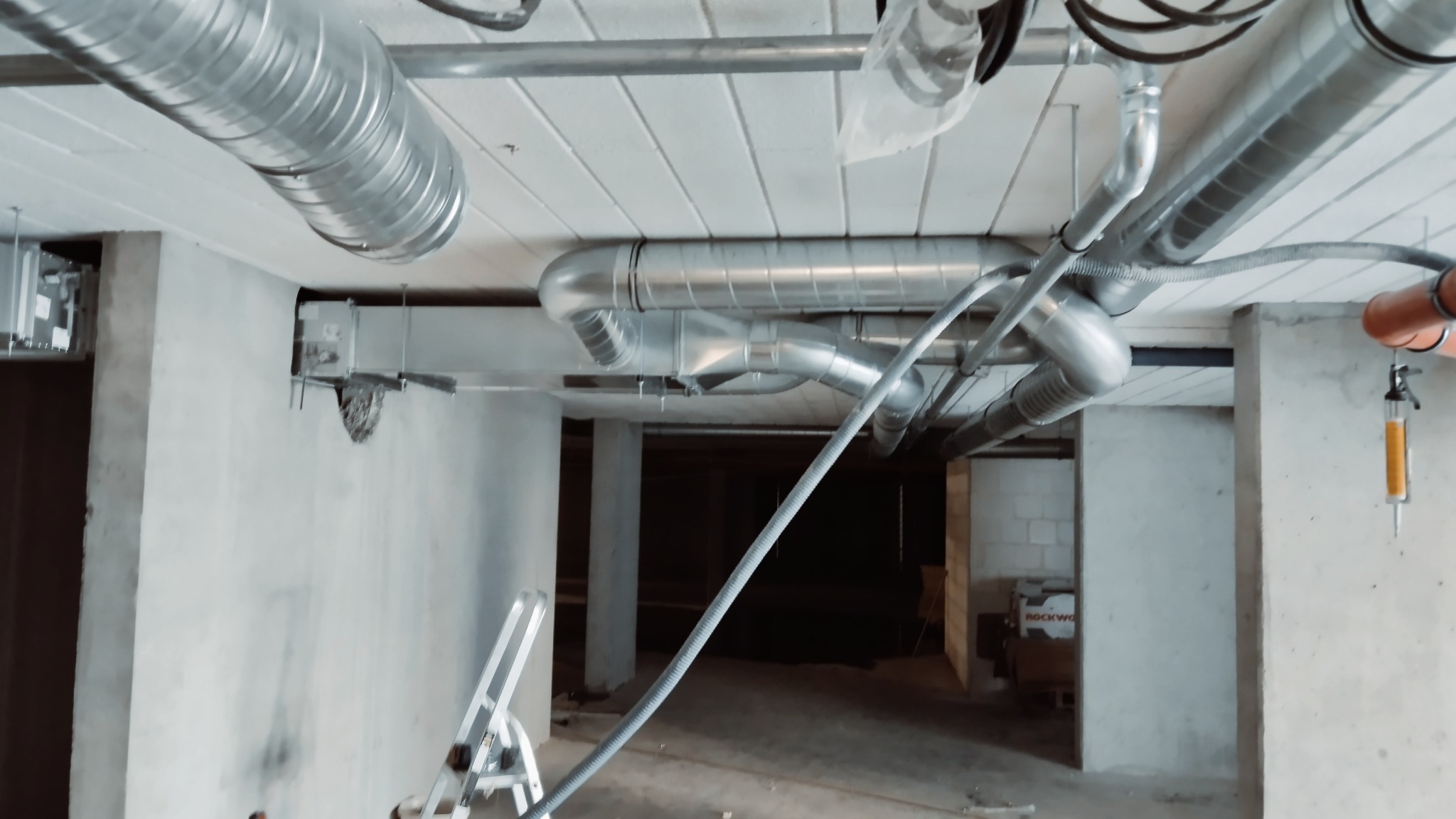 Widok instalacji wentylacyjnej z ocynkowanych rur i kanałów pod sufitem w surowym wnętrzu, drabina w tle, widoczne elementy mocujące i izolacyjne.