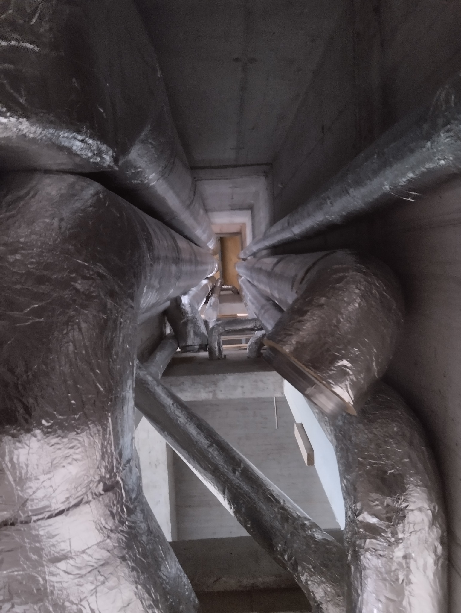 Wysoki kadr z dołu na system kanałów wentylacyjnych owiniętych srebrną folią izolacyjną, biegnących wzdłuż betonowego szybu wentylacyjnego w budynku.