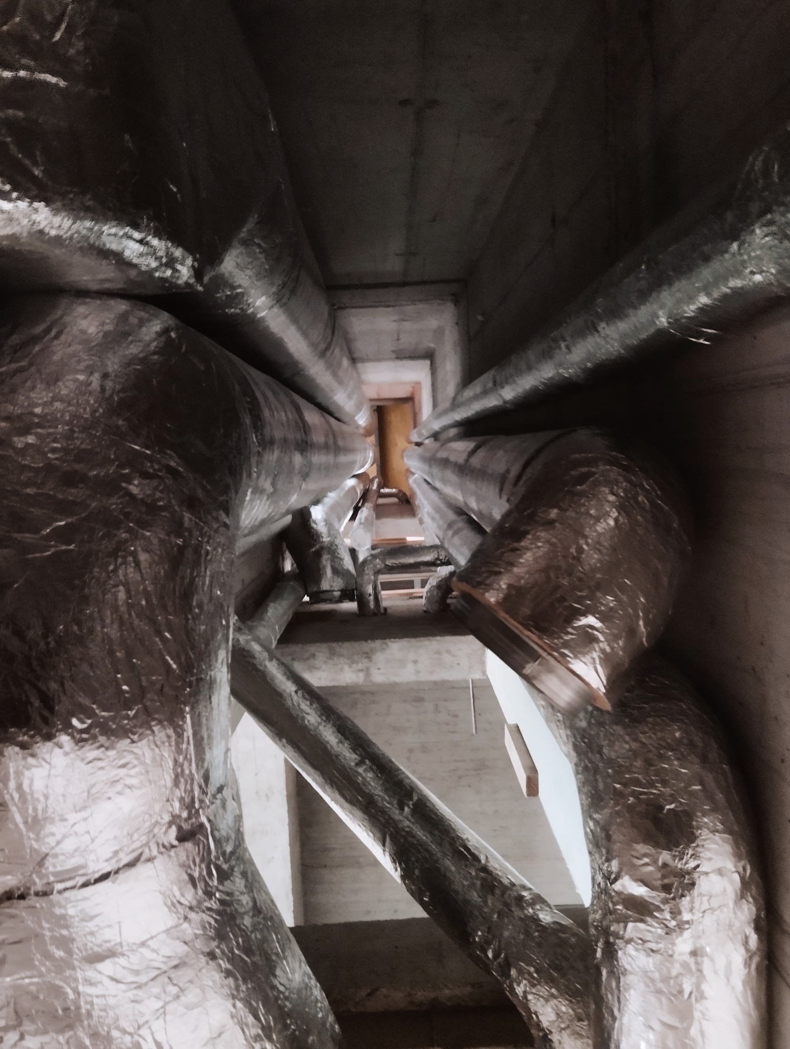 Wysoka perspektywa na system rur wentylacyjnych owiniętych folią izolacyjną w surowym, betonowym szybie wentylacyjnym.