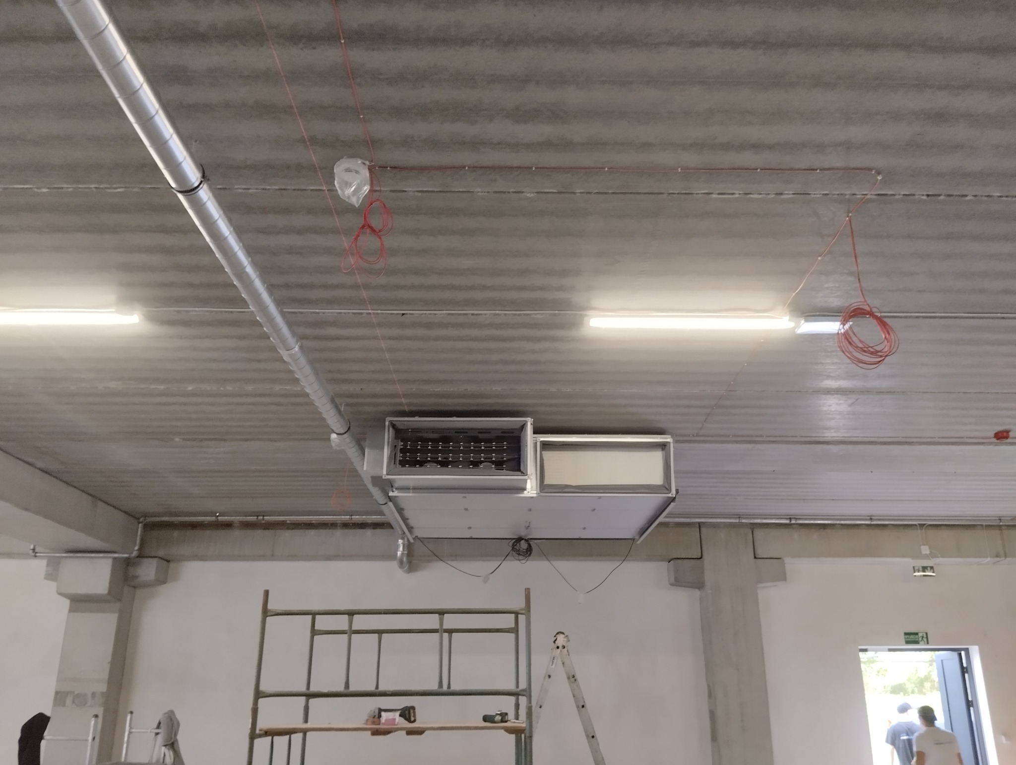 Montaż kanałów wentylacyjnych i rekuperatora pod betonowym sufitem w hali, widoczne kable i elementy instalacji, w tle rusztowanie i drabina