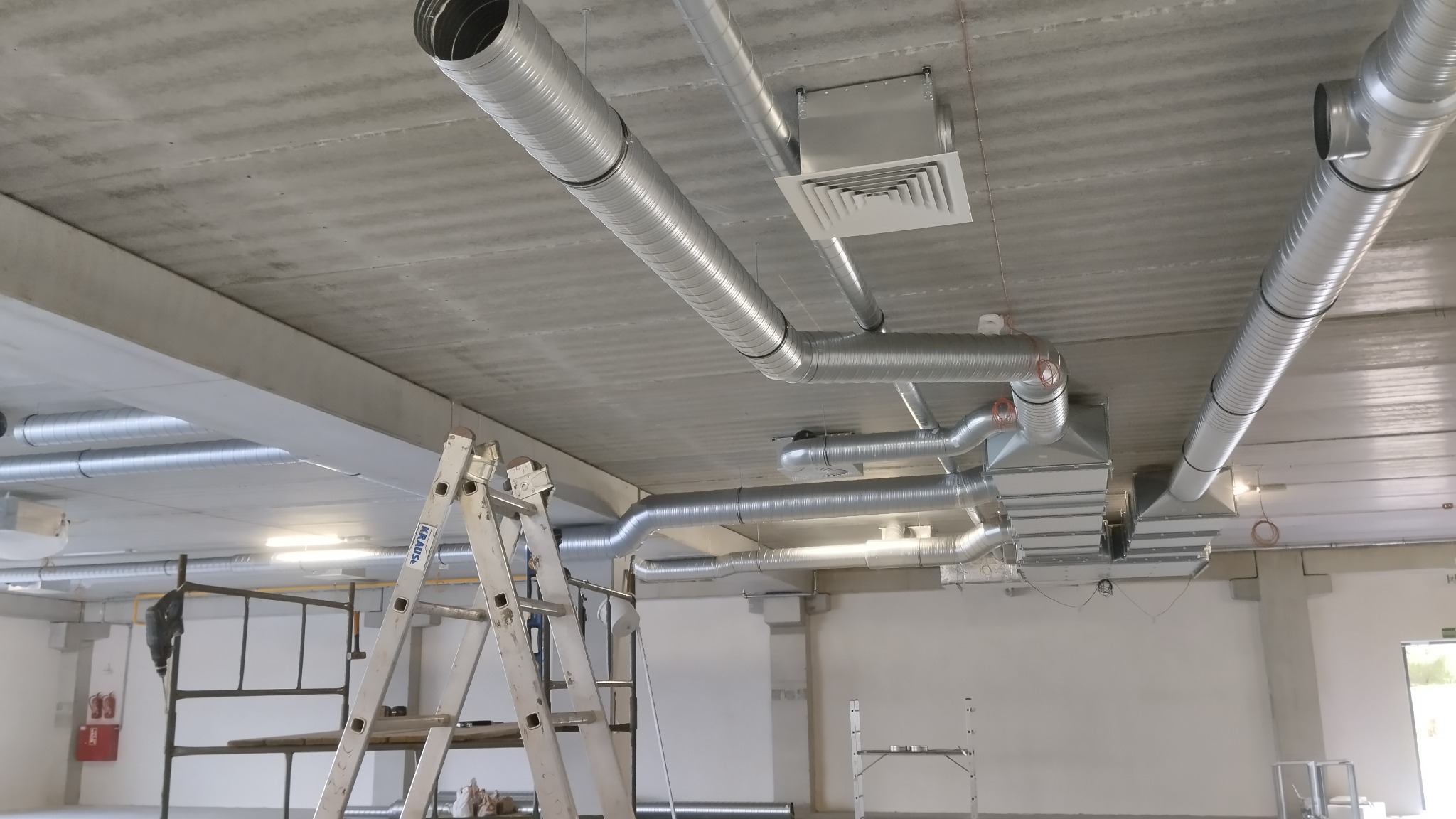 Srebrne, spiralne rury wentylacyjne zamontowane pod betonowym sufitem, widoczne drabiny monterskie i elementy instalacji, surowy stan wykończenia wnętrza.