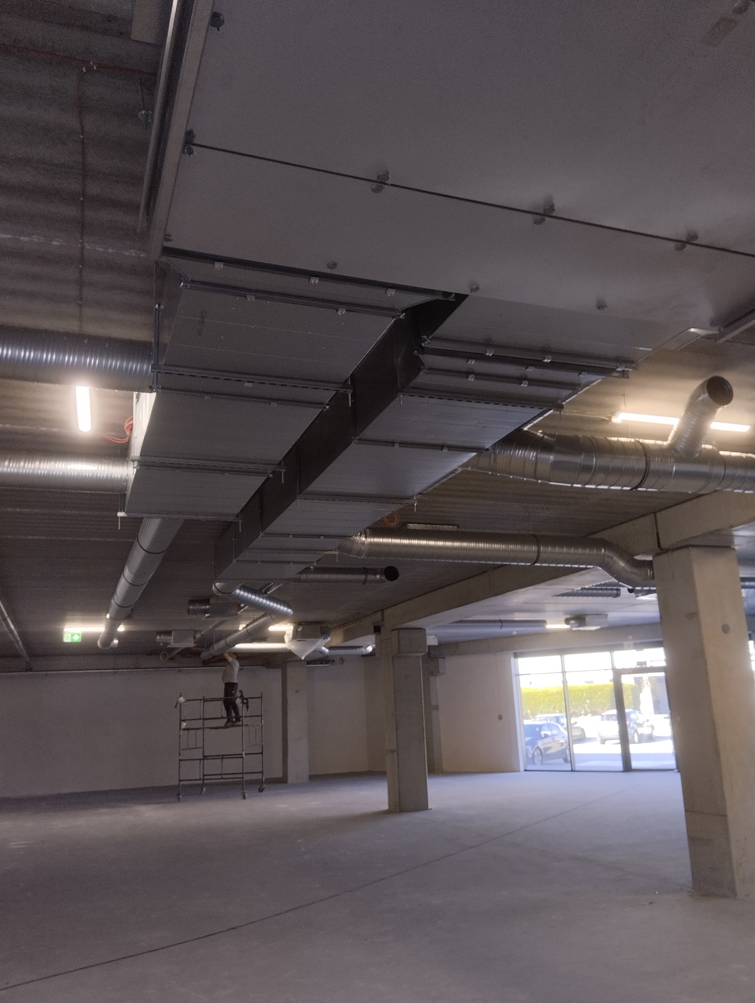 Sufit w hali przemysłowej z widocznymi kanałami wentylacyjnymi, rurami i pracownikiem na rusztowaniu podczas montażu.