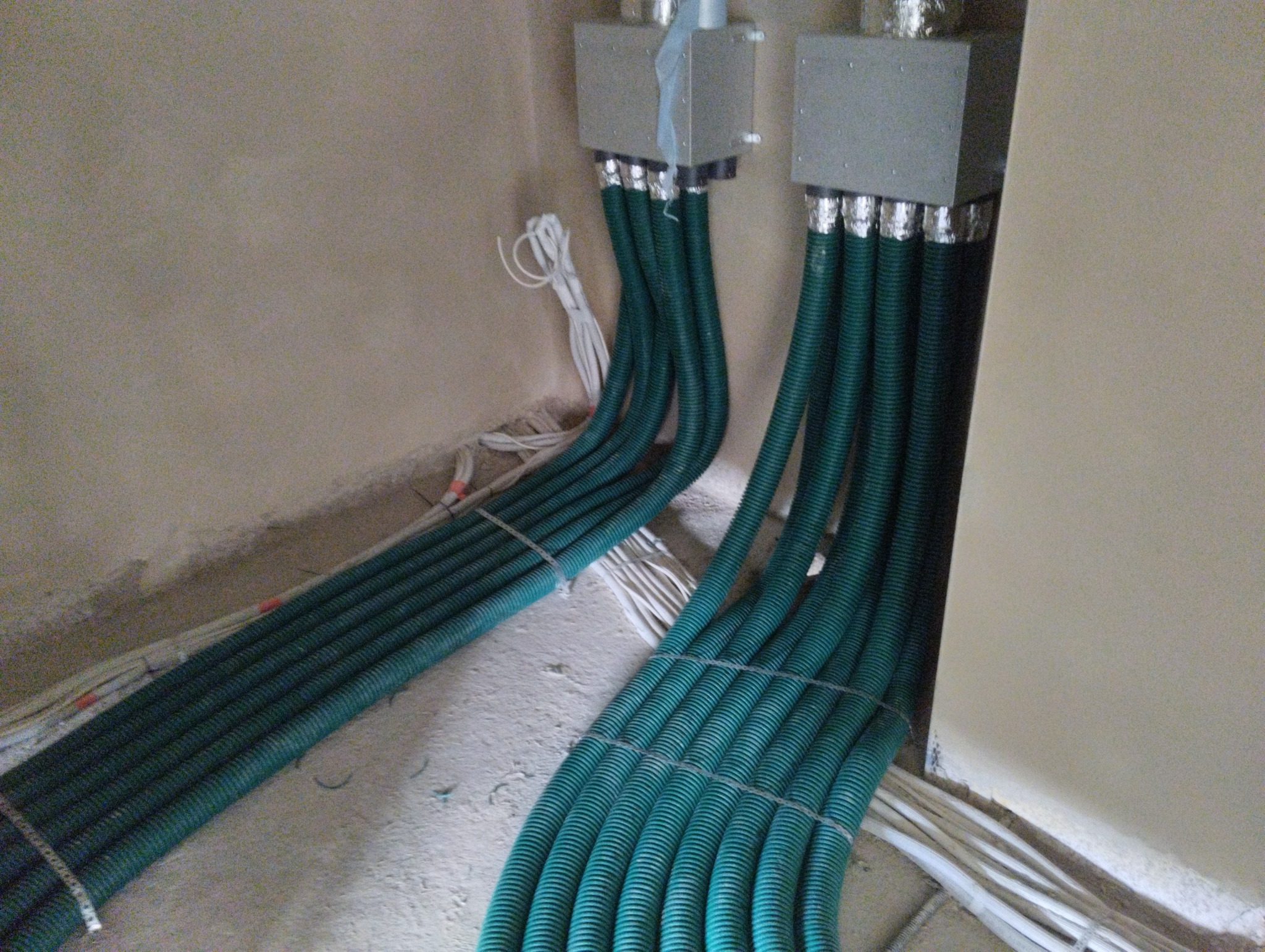 Szczegółowe ujęcie instalacji przewodów wentylacyjnych w budynku w trakcie montażu, widoczne zielone karbowane rury prowadzące do metalowych skrzynek na ścianie, z kablami elektrycznymi w tle.