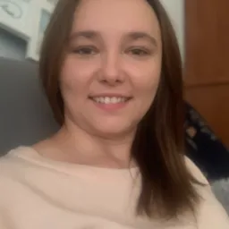 Portret uśmiechniętej kobiety z brązowymi włosami na neutralnym tle, prezentujący pozytywne nastawienie.