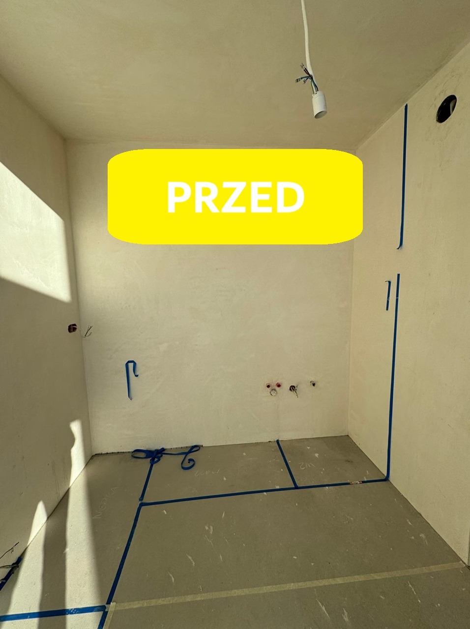 Pusty pokój przed remontem z widocznymi śladami po instalacjach, zabezpieczony niebieską taśmą malarską na podłodze i ścianach, z wiszącą żarówką.