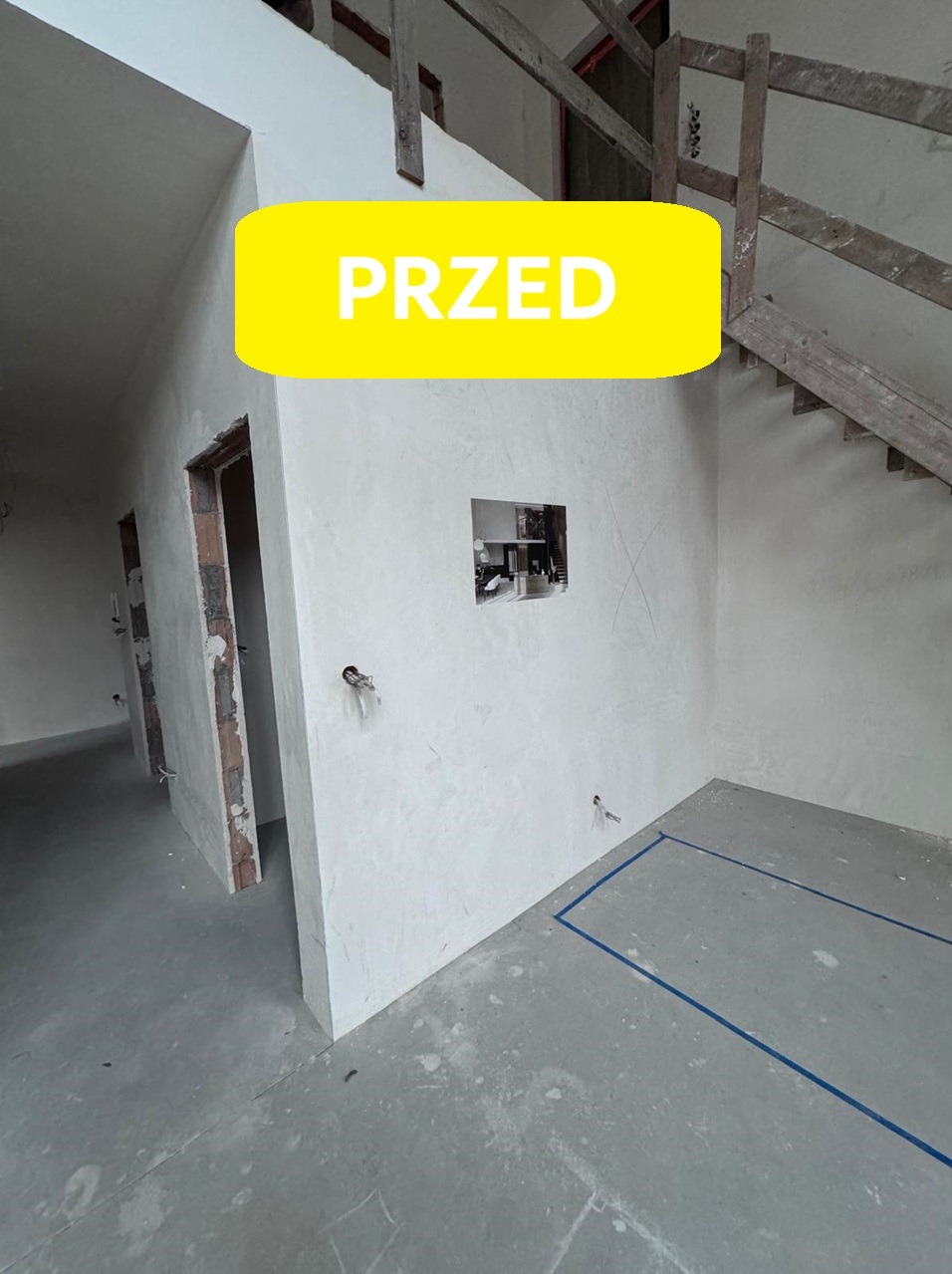 Wnętrze w trakcie remontu: surowe ściany, odsłonięte cegły wokół ościeżnic, nieotynkowana ściana z otworem, surowy beton na podłodze z niebieskim obrysem, drewniane schody w tle, naklejka z napisem...