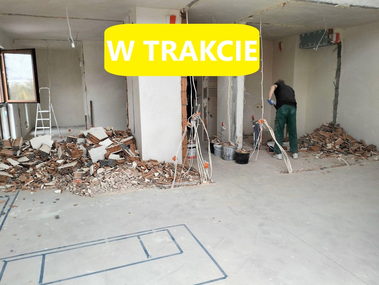 Demontaż ściany w trakcie: widoczne gruzowisko, odsłonięte przewody elektryczne, zarys planu pomieszczenia na podłodze oraz pracownik w tle.