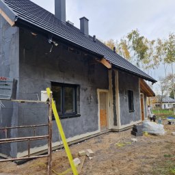 Zakład ogolnobudowlany Rzepka Tomasz Stanisław - Dom w trakcie ocieplania elewacji styropianem grafitowym, widoczne rusztowania, paczki styropianu Styropian Plus, przygotowane wejście do budynku, teren budowy z piaskiem i częściowo ułożoną kostką.